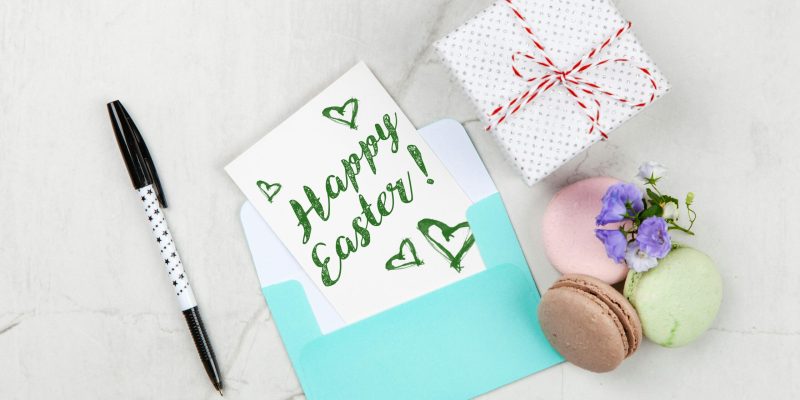 Flat lay of Easter greeting card with gift box, macarons, flowers, and pen on marble background.